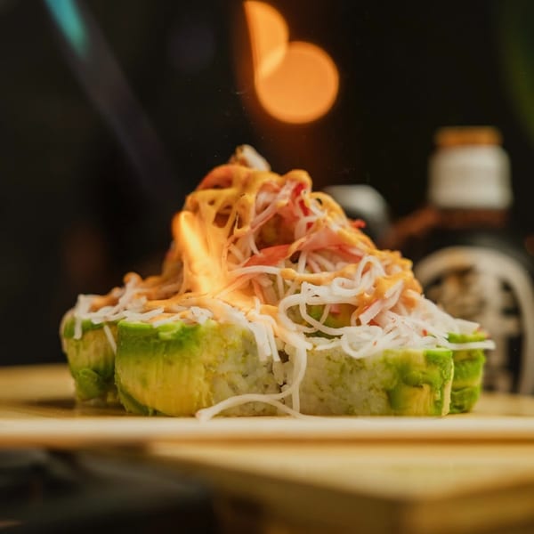 Flaming sushi rolls topped with crab meat, avocado, and spicy sauce on a wooden board.