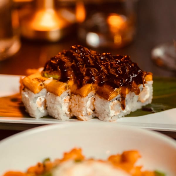 Sushi rolls topped with mango and a dark sauce, served on a white plate with warm lighting.