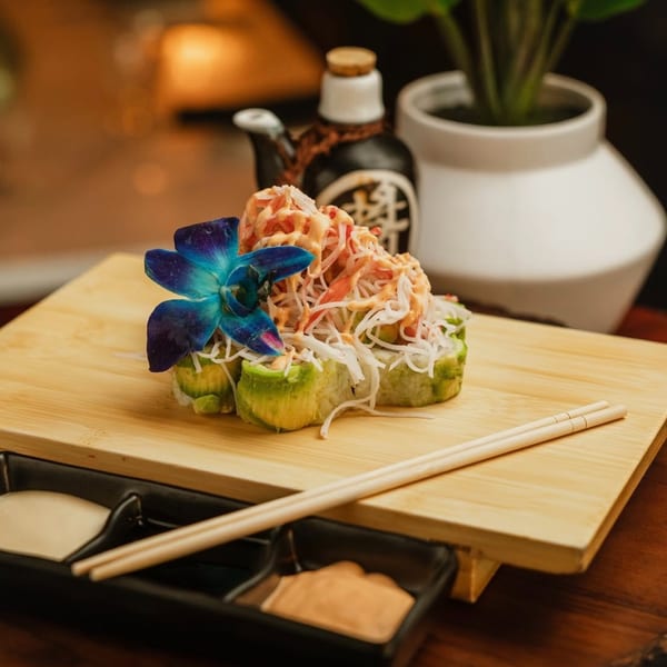 Sushi rolls topped with crab and sauce, garnished with a blue orchid on a wooden board.