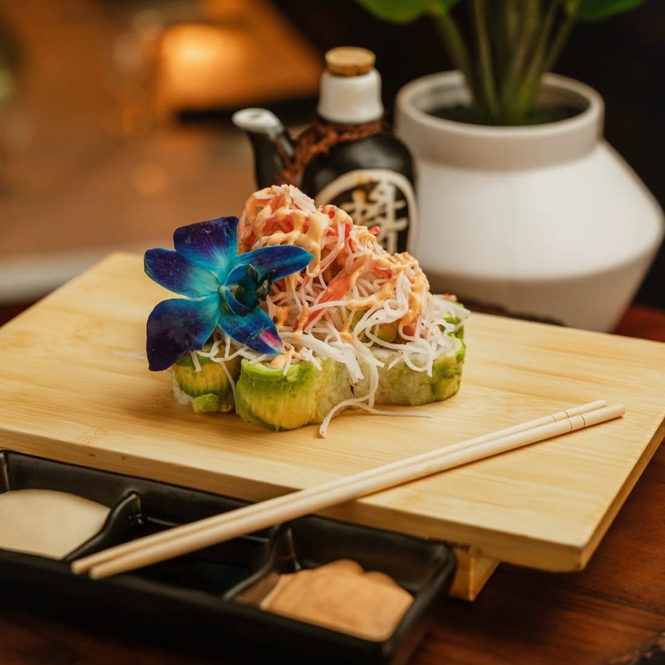 Sushi rolls topped with crab and sauce, garnished with a blue orchid on a wooden board.