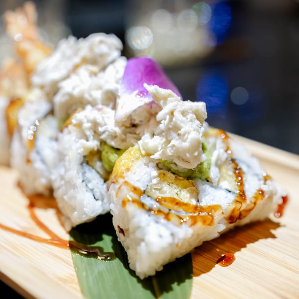 Sushi rolls topped with white crab meat, avocado, and a purple orchid on a wooden board.