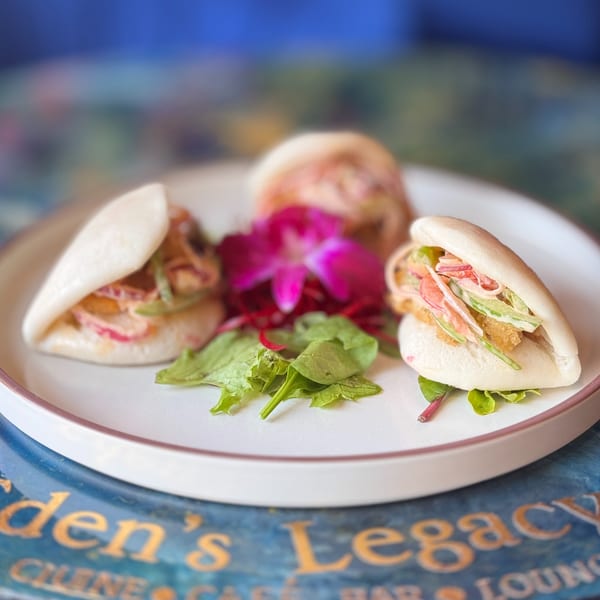 Three Dominican bao buns with crispy chicken, slaw, and sauce, garnished with a pink orchid.