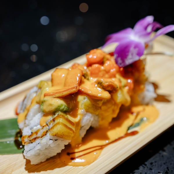 Sushi roll topped with orange sauce, shrimp, and a purple orchid on a wooden board.