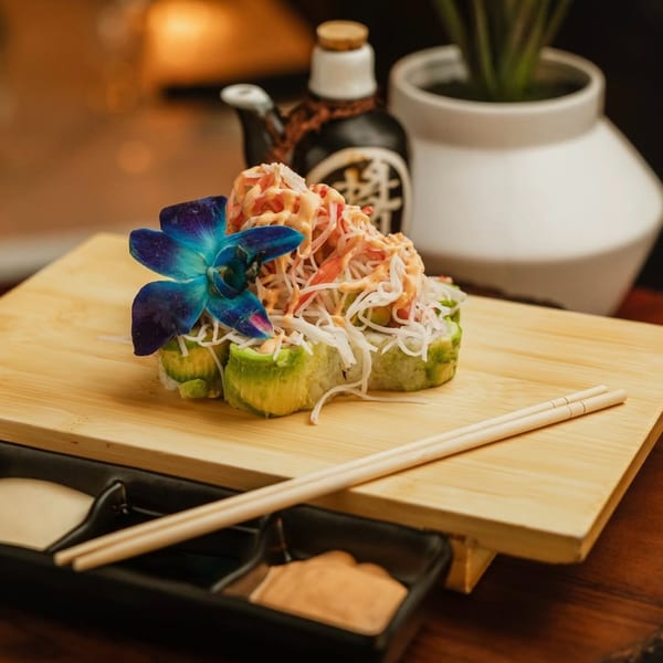 Sushi rolls topped with crab salad and a blue orchid, served on a wooden board with chopsticks.