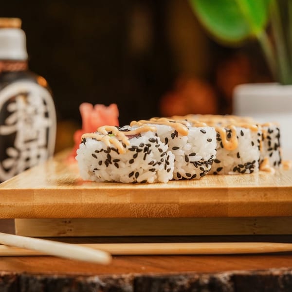 Sushi rolls with black sesame seeds and a creamy sauce on a wooden board with chopsticks.