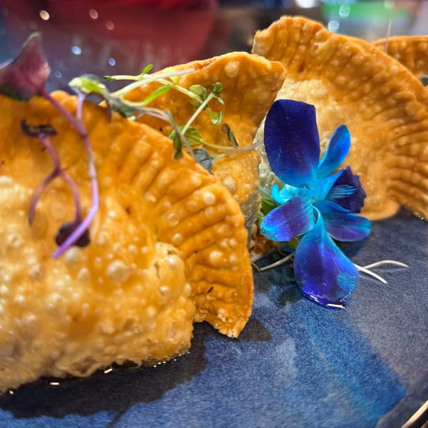 Golden-fried Dominican empanadas garnished with microgreens and a vibrant blue orchid on a dark plate.