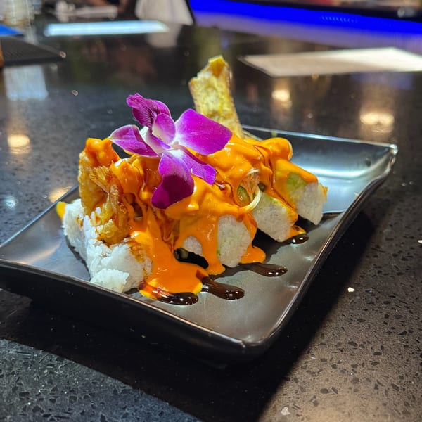 Sushi rolls topped with orange sauce, a purple orchid, and a crispy spring roll on a black plate.