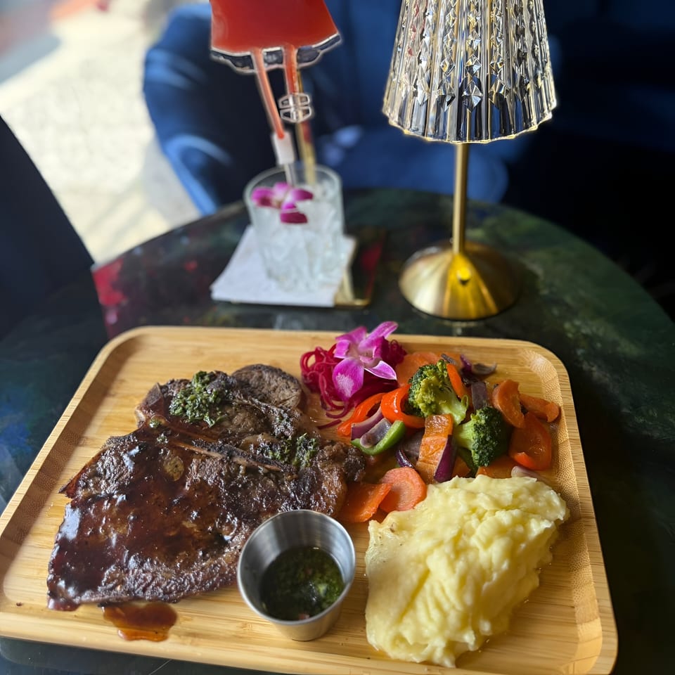 Dominican steak with mashed potatoes, mixed vegetables, and a cocktail on a wooden tray.
