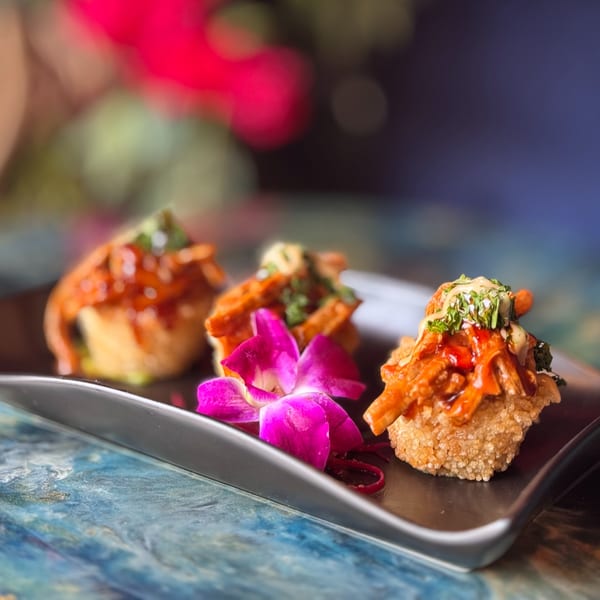 Three Dominican appetizers with sauce and herbs, garnished with a purple orchid on a black plate.