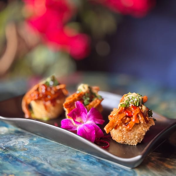 Three crispy Dominican appetizers topped with sauce, herbs, and a pink orchid on a black plate.