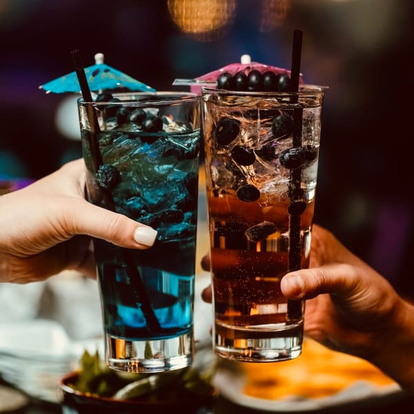 Two hands holding tall glasses of blue and amber cocktails with blueberries and umbrellas, cheering.