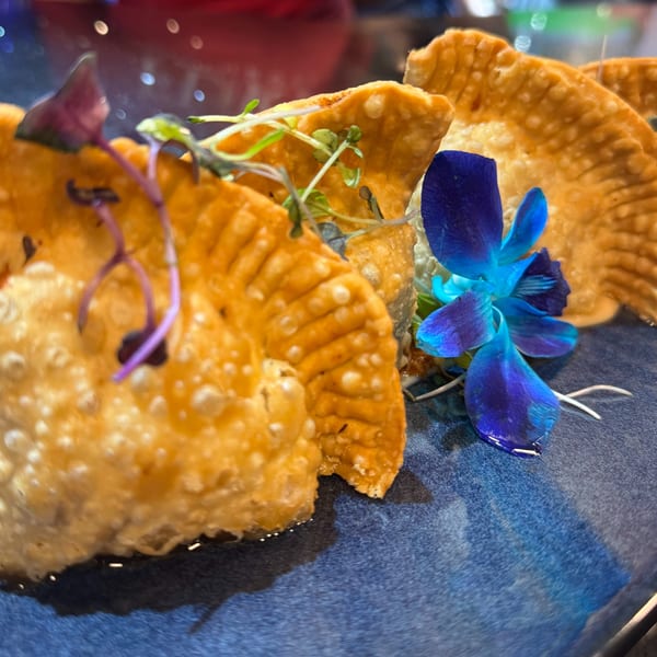Golden-brown Dominican empanadas garnished with microgreens and a vibrant blue orchid on a dark plate.