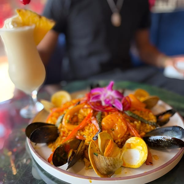 Dominican seafood paella with mussels, clams, shrimp, and vegetables, garnished with an orchid, next to a pina colada.