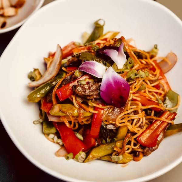 A white bowl filled with Dominican noodles, red and green peppers, onions, and purple orchid petals.
