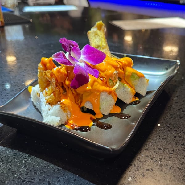Sushi rolls with orange sauce, a purple orchid, and a fried spring roll on a black plate.