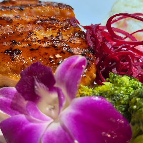 Close-up of glazed Dominican pork, red pickled onions, broccoli, and a purple orchid garnish.